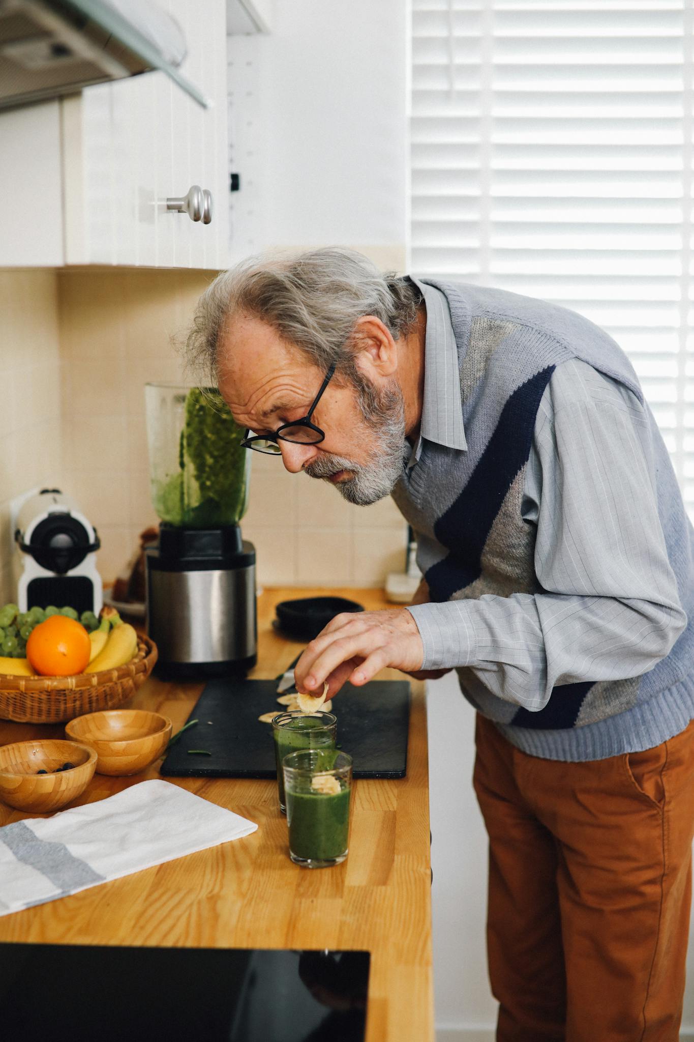 Elderly man making a healthy green smoothie in a kitchen with fresh fruits.